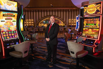 Older white man with receding gray hair wearing a black pinstripe suit, white collared shirt, red tie and gold watch on his left wrist poses with his hands crossed in front of him while standing in a casino and flanked by two slot machines