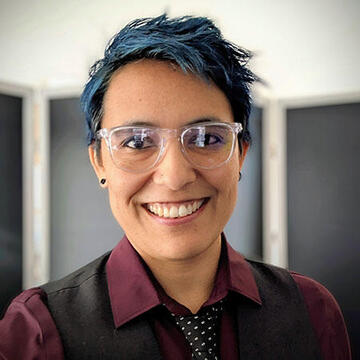 Headshot image of a smiling Latin woman with short dark hair with blue highlights wearing clear eye glasses, stud earrings and a burgundy-colored collared shirt