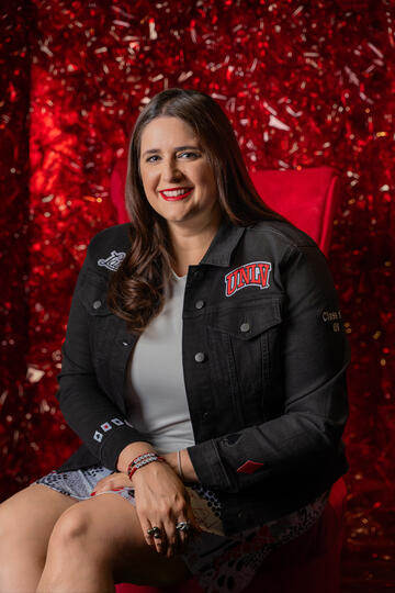 White woman with long brown hair wearing a dark UNLV jacket and gray top smiles while sitting on a bright red chair against a bright red background