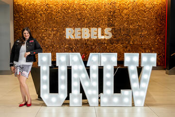 White woman with long brown hair wearing a dark UNLV jacket, gray top, red, white and black skirt, and red shoes smiles while posing with her left elbow on an oversize UNLV sign and in front of a gold wall that has the word Rebels illuminated in white on the wall 