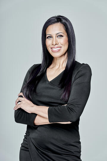 Dark-complected woman with long straight black hair wearing a black dress smiles as she poses with her arms crossed in front of her