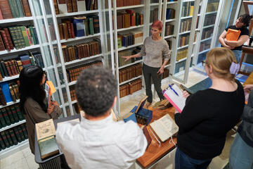 woman speaking to a group of people in front of a wall of bookcases filled with older books.
