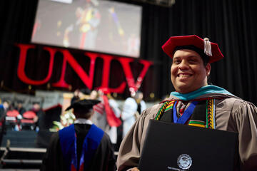 Greenspun College of Urban Affairs master's graduate holds his degree in front of the UNLV lights.