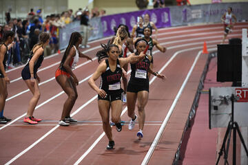 A black female wearing a UNLV track jersey reaches back with her left hand to take a baton from another black female UNLV sprinter during a collegiate track and field race