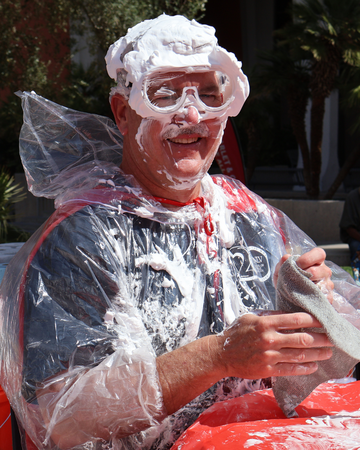 White male wearing a plastic poncho and goggles smiles after getting hit with a whipped cream pie 
