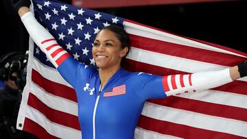 A Black woman in a Team USA bobsled suit smiles and extends her arms as she holds an American flag behind her