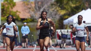 A young Black woman in a UNLV jersey sprints down the track with two other Black sprinters running on both sides of her