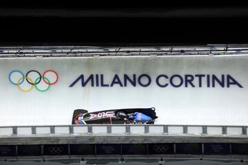Image of a blue bobsled racing down a bobsled track with the Olympic rings and the words Milano Cortina displayed on the track above the bobsled