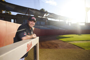 White man wearing a black UNLV baseball cap and short-sleeve black UNLV pullover smiles as he leans over the rail of a baseball dugout at UNLV's baseball field while bright rays of sun shine in the background 