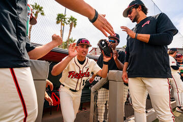Baseball player in a UNLV Rebels No. 14 uniform with a glove on his left hand emerging from the dugout and high-fiving teammates