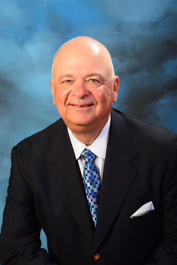Older white man with a bald head wearing a dark blue suit, white collared shirt and multicolored tie with a white hankie partially sticking out of his suit pocket poses against a blueish background
