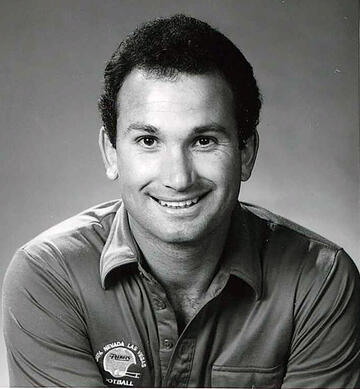 Vintage black and white photo of a young white male with black hair and a dark polo shirt with a UNLV football logo on the left breast smiles while posing against a gray background