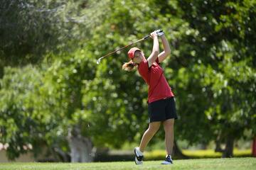 White female golfer in a red top, black shorts, white shoes and red hat with a pony tail follows through on a golf swing