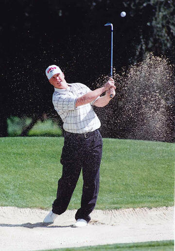 Sand hangs in the air after a white man wearing dark pants, white plaid shirt, white shoes and a white hat with a red UNLV logo follows through on a golf swing after hitting a ball out of the sand trap on a golf course 