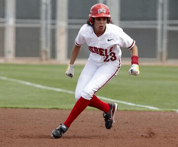 White female softball player wearing an all-white uniform with the word Rebels and No. 23 in red on her jersey and a red UNLV helmet with red socks, black cleats, red wristbands and white batting gloves prepares to steal a base on the dirt infield of a softball field