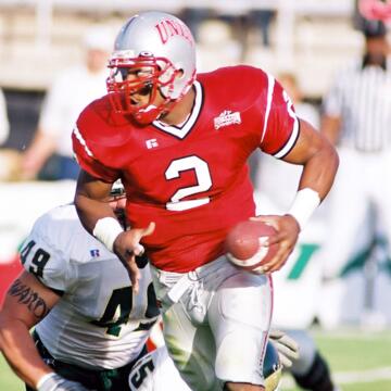 Black football player in a red No. 2 jersey, silver UNLV helmet with red facemask and gray pants with a red stripe down the side carries a football in his left hand while running away from a partially obscured opponent who is wearing a white jersey with the No. 49 in black