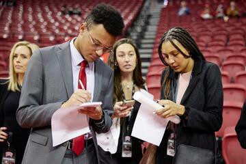 Three students write on notepads while covering a basketball game for a sports broadcast.