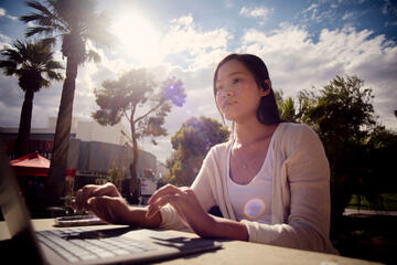 A student studies at a laptop in an outdoor seating area in front of palm trees as sun shines through the clouds