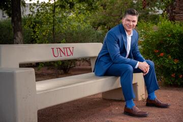White man in a royal blue suit and brown loafers leans forward with his elbows on his knees while sitting outside on a bench with the UNLV logo