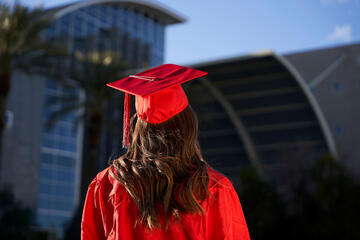 a female student with dark hair and wearing a red cap and gown is shown from the rear, facing Lied Library