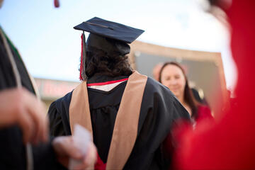 The back of a student graduating with their master's degree, wearing the graduation cap and gown and a light brown stole.
