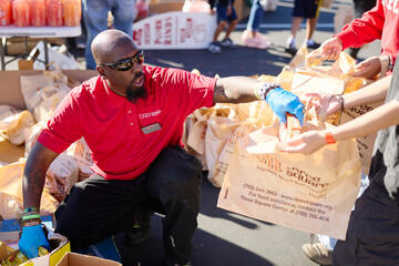 A man wearing a red UNLV polo places food into a plastic bag bearing a Three Square logo