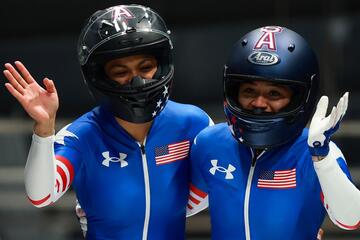 Two female bobsled competitors wearing blue Team USA skin suits and helmets wave to the camera