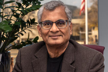 man in jacket posing iman in jacket posing in an office with image of buildings and flag visible behind himn an office with buildings and flag visible through window.