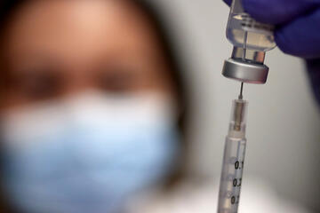 A masked medical worker inserts a syringe into a vial containing COVID vaccine solution