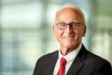 Older man with short gray hair smiles as he poses against a blurred background while wearing a white collared shirt, black suit jacket, red checkered tie and eyeglasses