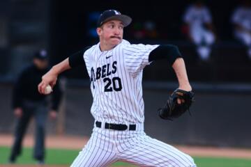 White male baseball pitcher in a black and white pinstripe Rebels uniform with the No. 20, black stirrup socks and a black baseball cap grips a baseball in his right hand, reaches behind himself and prepares to deliver a pitch