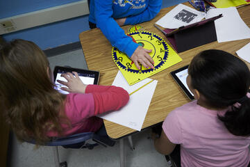 Two elementary school students and their principal sit around a desk with tablets, paper, and pencils