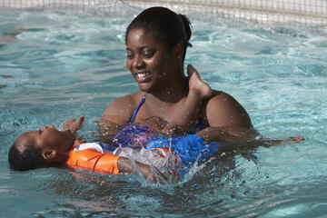 smiling woman in pool holds toddler as he practices swimming on back
