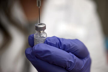 medical professional pokes syringe into top of a vial she holds in her gloved left hand