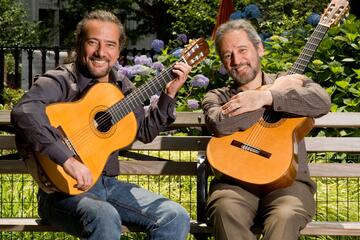Sérgio and Odair Assad, holding their guitars, look towards the camera. They are in a springlike setting.