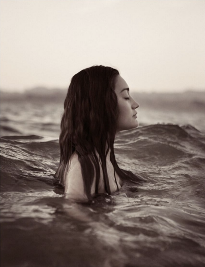 A woman with long dark hair stands in profile, chest deep in wave-filled waters. Her eyes are closed, her face serene.