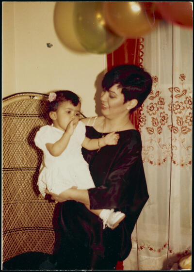 Young one-year old child in a white dress being held by her mother with short hair and a black shirt. There are gold balloons and strong shadows from a flash.