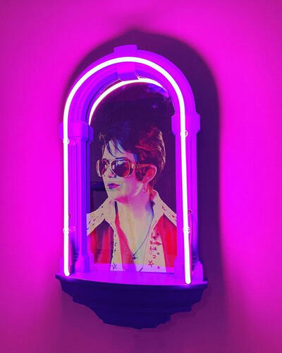 An alter-like object hangs on a wall. A neon tube curving around the top of the altar throws purple-pink light over the entire picture. The interior of the altar is filled with the image of someone dressed as Elvis, with sunglasses and a black pompadour.