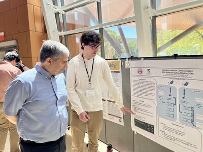 A student who participated in the REU experience points to his research poster while his mentor looks on