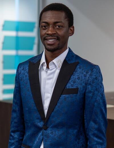 A portrait of Nehemiah Afam Chukwuere wearing a blue blazer and standing in a conference room