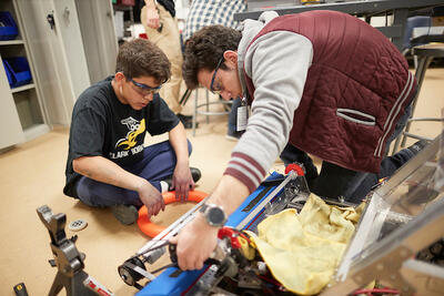 Middle school boy sitting on the ground and bulding a robot with an adult assisting him.