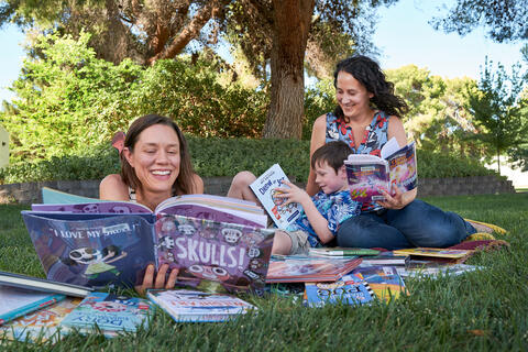 two women and child reading books on a blanket outdoors