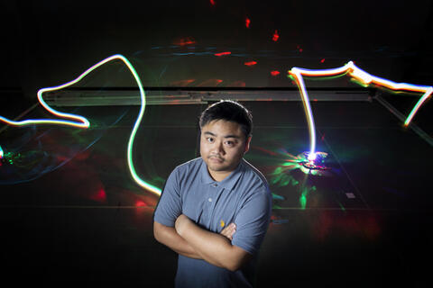 male student with arms folded and streaks of light from aerial drones in the background