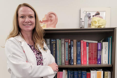 A woman wearing a white labcoat stands in front of a bookshelf loaded with books and a model of an ear.