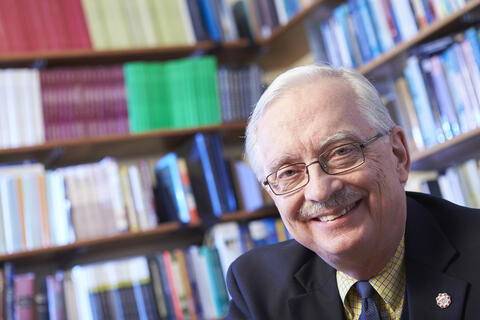 Stephen M. Miller smiling and sitting behind a shelf filled with books