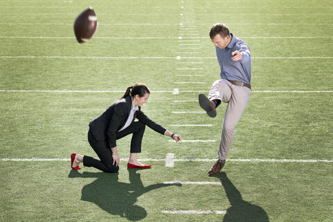 Evan Pantels kicks a football as Mallory Pool holds it.