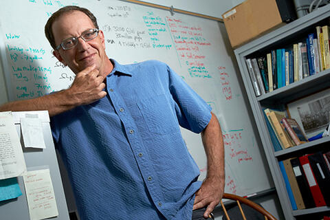 A man leaning against a file cabinet with a white board and a shelf filled with books behind them