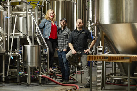 a woman and two men surrounded by brewery equipment