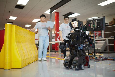 Avatar-Hubo gets ready to stand as two students work on computers.