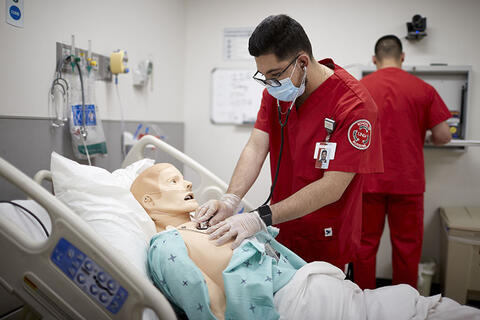 Two nursing students in a simulation, one is in the background working on something unseen, the other is using a stethoscope on a dummy patient on a hospital bed 